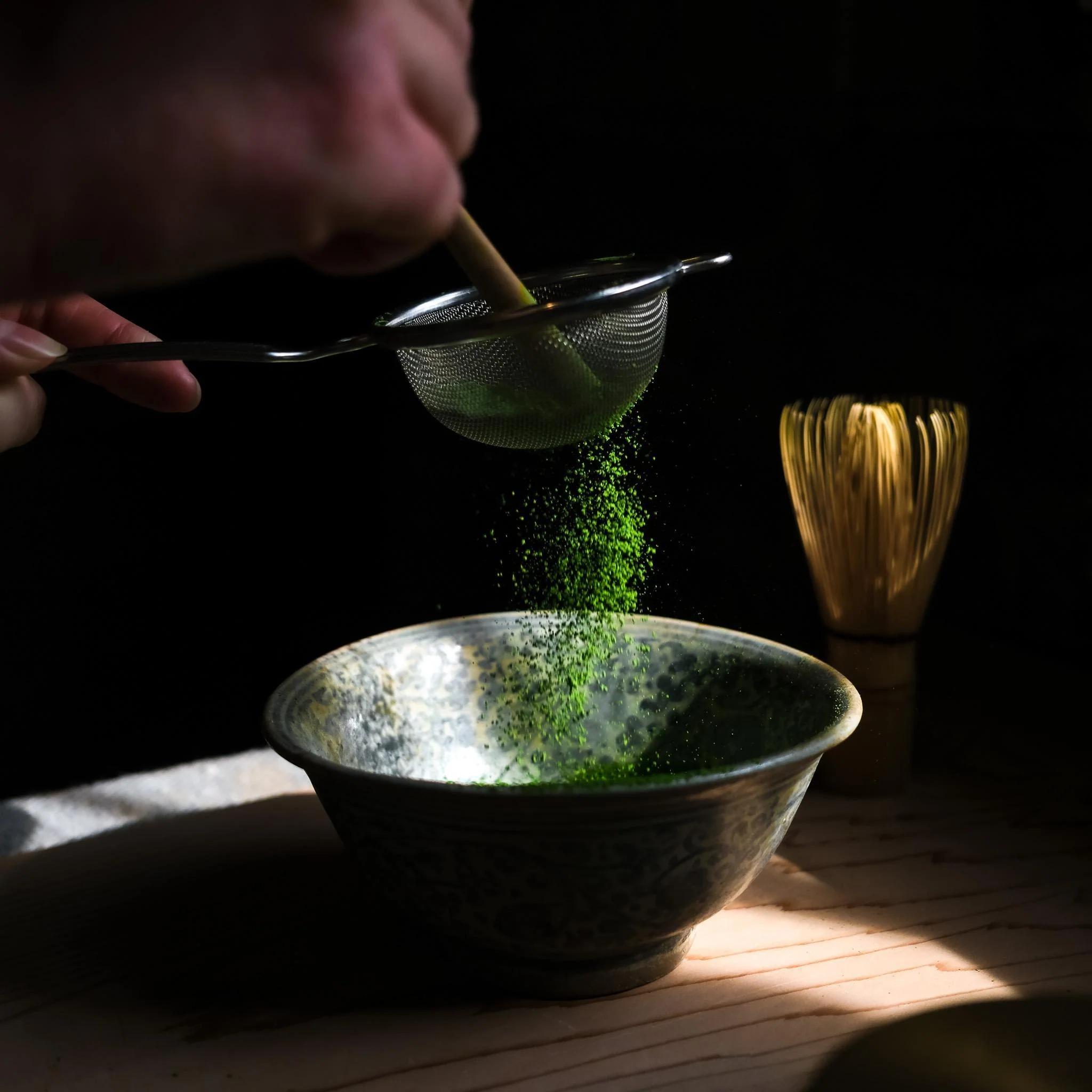 Matcha powder being sifted through a fine mesh strainer into a traditional ceramic bowl, highlighting its micro-fine particle size and vibrant green color.